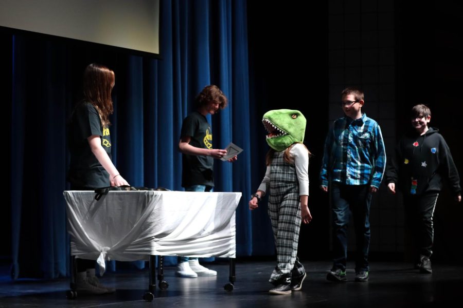 A student in a dinosaur head piece receives her certificate for her video, A Very Heartwarming Tale about a Very Dramatic Dinosaur Finding a Name. 