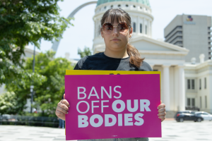 Senior Athena Widlacki stands in front of the St. Louis Arch on Kiener Plaza, with one of the free #BansOffOurBodies posters.