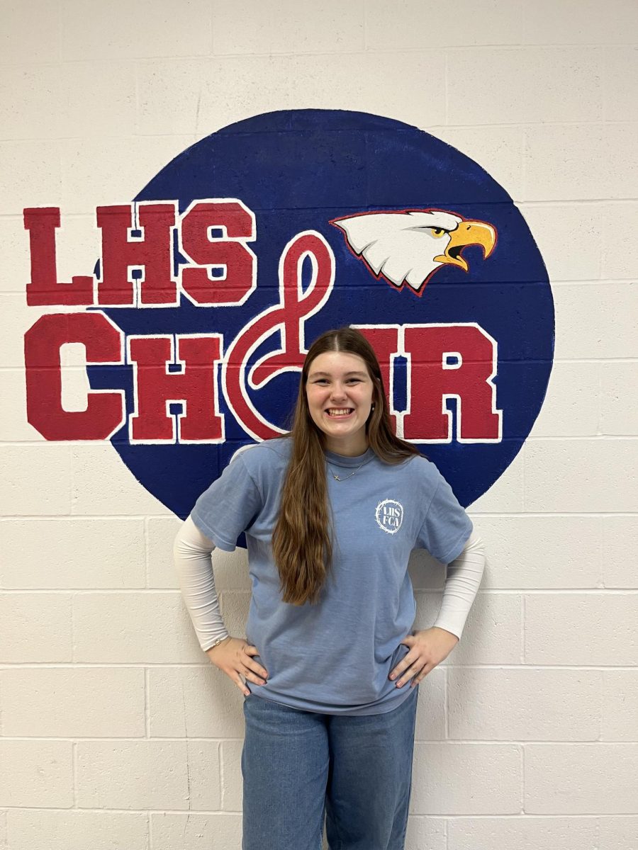 Ellie Force (11) stands proudly in front of the LHS Choir logo in the 500 hallway.