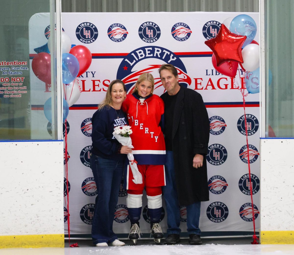 Diana Kemp (12) poses with her mom and dad in front of Liberty Hockey backdrop for senior night.