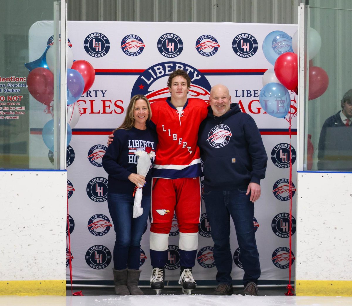 Zach Pruss (12) poses with his mom and dad in front of Liberty Hockey backdrop for senior night.