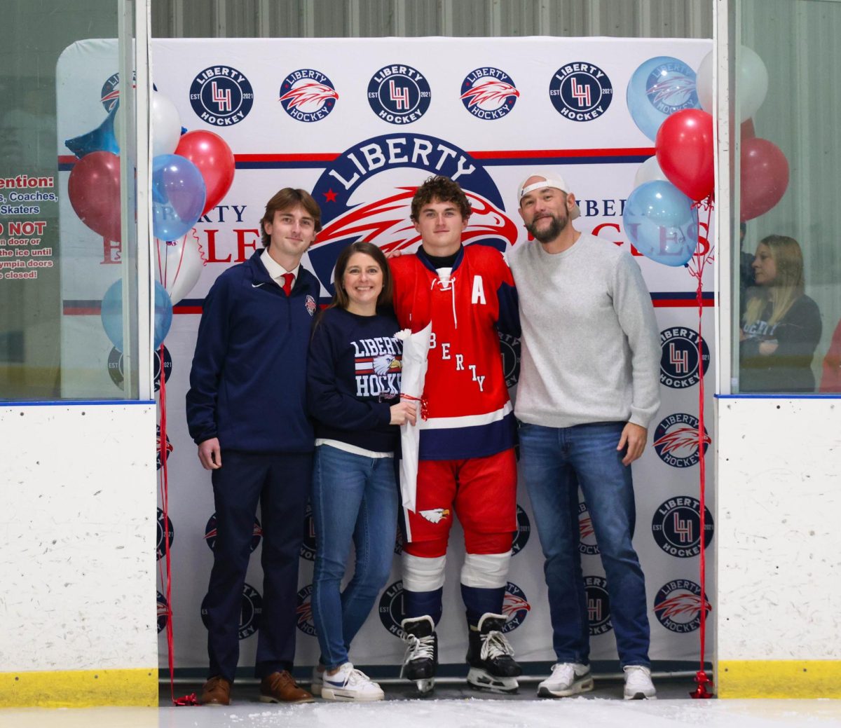 Greyson Howarth (12) poses with his brother, mom, and dad in front of Liberty Hockey backdrop for senior night.